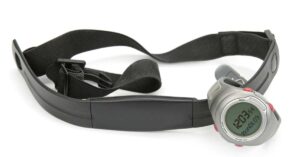 A digital watch with a black strap and a metal case is displayed on a white background. The watch is attached to a dark, adjustable chest strap heart rate monitor. The watch screen shows a time of 12:03:54.