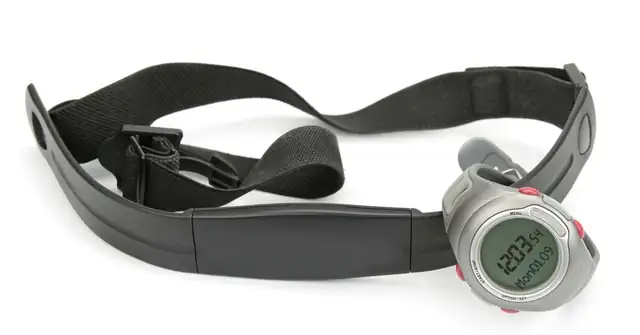 A digital watch with a black strap and a metal case is displayed on a white background. The watch is attached to a dark, adjustable chest strap heart rate monitor. The watch screen shows a time of 12:03:54.