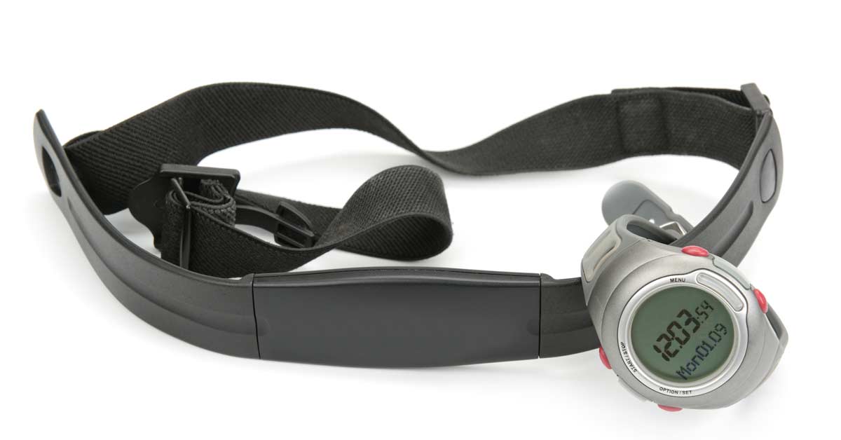 A digital watch with a black strap and a metal case is displayed on a white background. The watch is attached to a dark, adjustable chest strap heart rate monitor. The watch screen shows a time of 12:03:54.