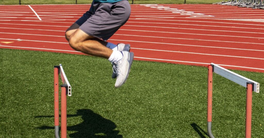 A person in athletic shorts and a blue shirt is seen mid-air, jumping over a hurdle on a track. The track is red with white lines, and the field is covered in green artificial turf.