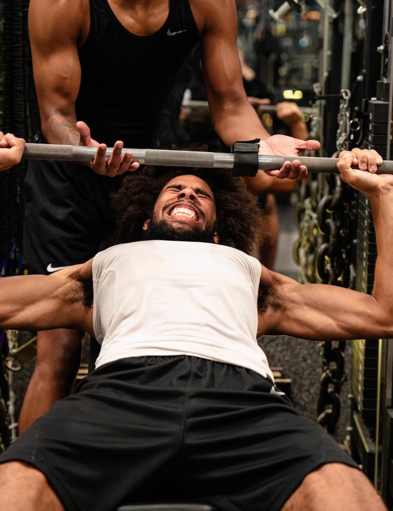 A man straining as he lifts a barbell while bench pressing at the gym, with another person standing behind him, spotting and assisting.