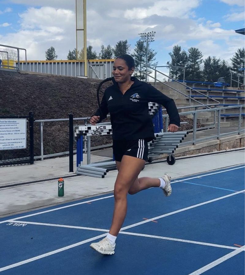 A woman in athletic wear runs on a blue outdoor track, smiling. Bleachers and trees are in the background, and a water bottle sits near the track. The sky is partly cloudy.
