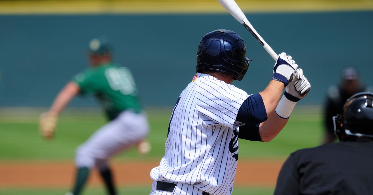 A baseball player in a white pinstripe uniform prepares to bat as a pitcher in a green jersey throws the ball. An umpire crouches behind the batter. The scene is set on a sunny outdoor baseball field.