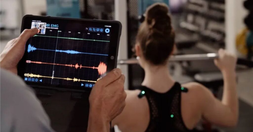 A person holds a tablet displaying colorful waveforms and graphs while another person, viewed from behind, lifts a barbell in a gym setting.