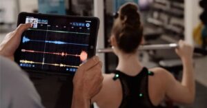 A person holds a tablet displaying colorful waveforms and graphs while another person, viewed from behind, lifts a barbell in a gym setting.