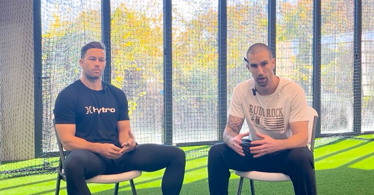 Two men sit on chairs in a brightly lit indoor sports facility with large windows and green turf. One wears a black Hytro shirt, the other a white BLDR RCK shirt, and both look toward the camera.
