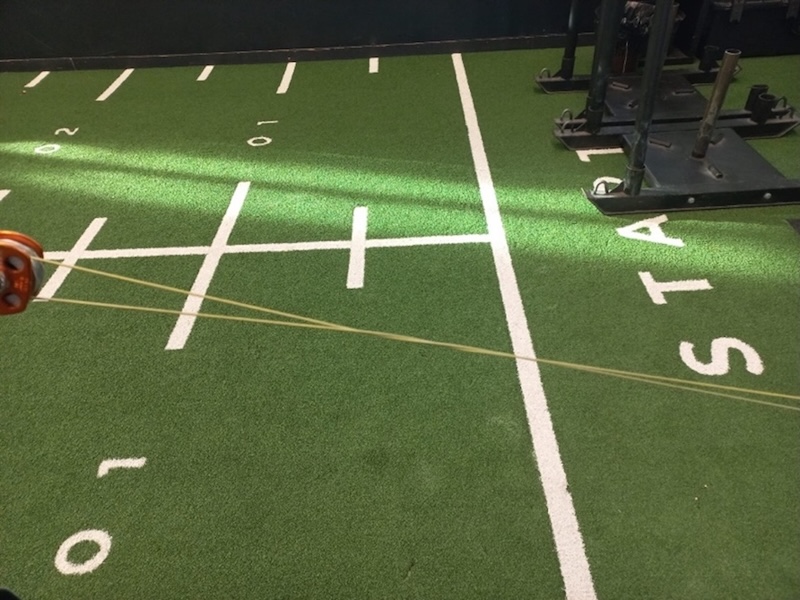 Green turf with white yard lines resembling a football field, gym equipment on the right, and a resistance band being pulled diagonally across the image from left to right.
