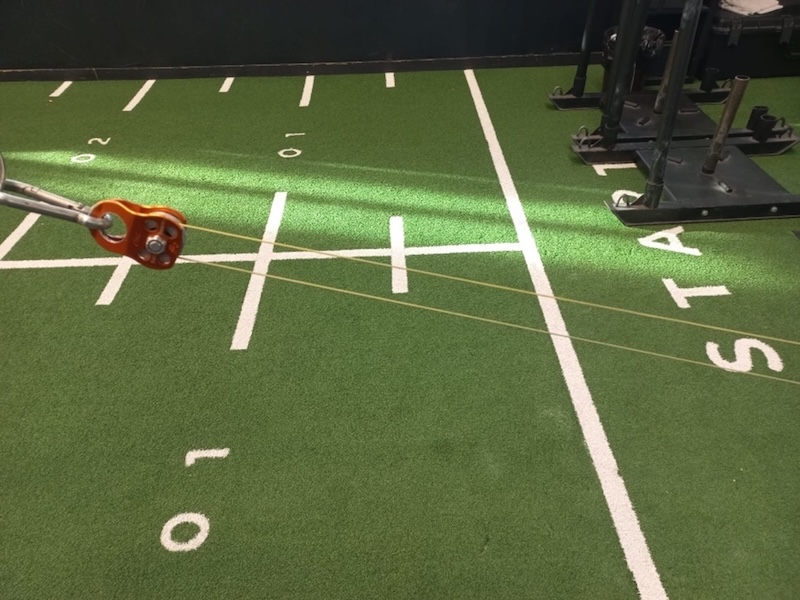Close-up of orange gym pulley with attached cables, positioned over green turf flooring marked with white yard lines and numbers, resembling a football field. Weightlifting equipment is visible in the upper right corner.