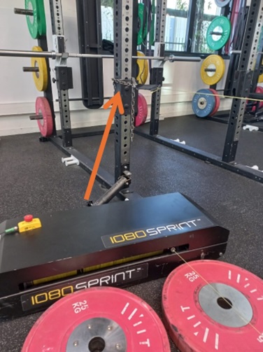 A gym squat rack with various colored weight plates is shown. An orange arrow points to a mechanism on the rack. In front, a black 1080 Sprint machine and pink 25 kg weight plates are visible on the floor.