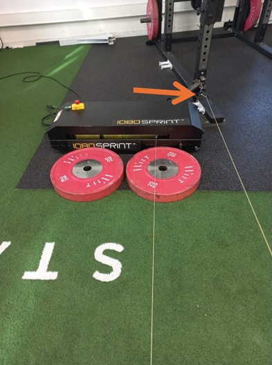 A 1080 Sprint training device with two red weight plates in front, set on artificial turf with white letters. An orange arrow points to a cable attachment on the device.