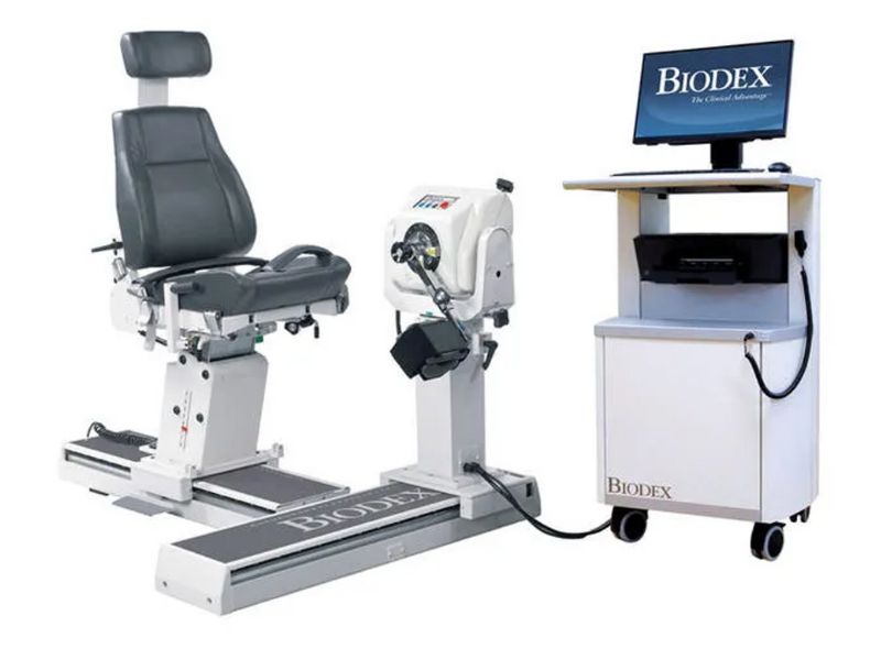 A Biodex dynamometer with a black seat, adjustable arm, and footrest. Its attached to a computer station with a monitor displaying the Biodex logo. The setup is used for physical therapy and rehabilitation assessments.