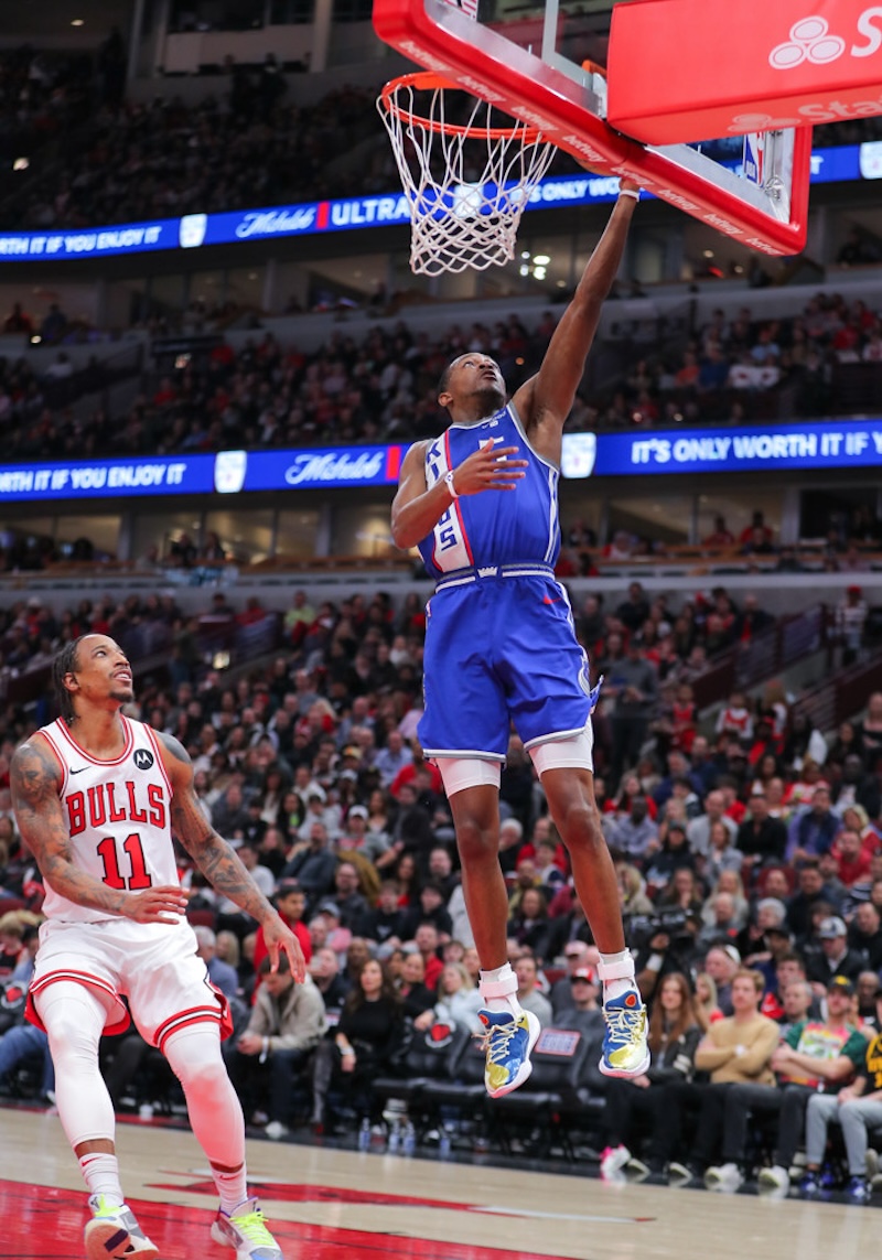 A basketball player in a blue jersey jumps towards the hoop for a layup, while another player in a white jersey watches. The packed stadium crowd and court are visible in the background.