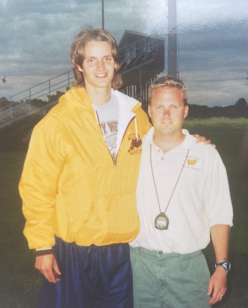 Two men posing outdoors on a grass field. The man on the left wears a yellow jacket and shorts, while the man on the right wears a white polo shirt and has a lanyard with a stopwatch around his neck. A sports stand is visible in the background.
