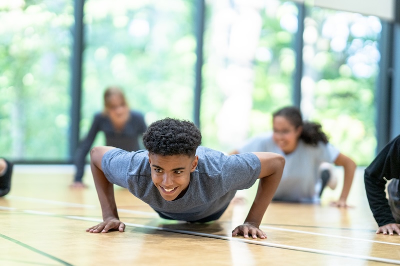 A group of people exercises indoors, focusing on a young person in a gray shirt doing push-ups on a wooden floor. A large window in the background offers a view of green trees, providing a bright and airy atmosphere.
