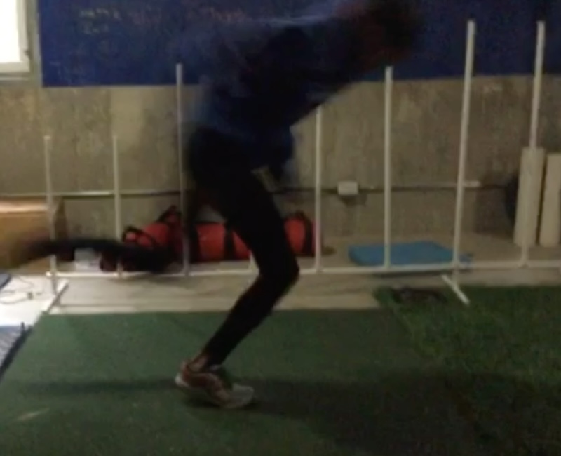 A person in motion, seemingly mid-jump or leaning forward, in an indoor setting. They are wearing athletic clothing and sneakers, with gym equipment and mats visible in the background. The image is slightly blurred.