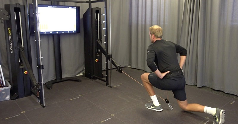 1080 Quantum|1080 Quantum Accessories|Pallof Horizontal Press|Overhead Pallof|A person in athletic wear performs a squat while holding a barbell overhead in a room equipped with fitness machines and a computer on a nearby desk. The area is carpeted