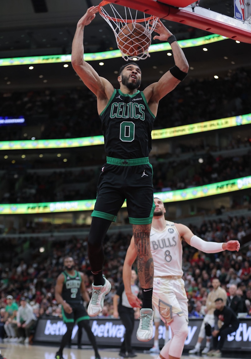 A basketball player in a black Celtics uniform jumps to dunk the ball during a game. Another player in a white Bulls uniform is visible in the background. The arena is filled with spectators.