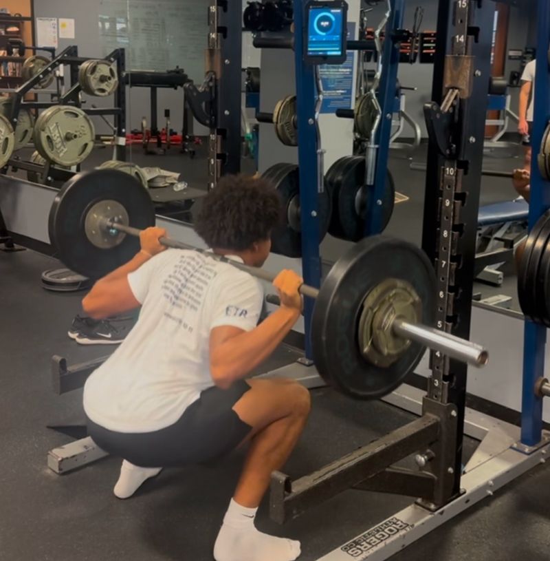 A person is performing a barbell squat in a gym, facing a squat rack. They are wearing a white T-shirt, black shorts, and white socks. Various gym equipment and weight plates are visible in the background.