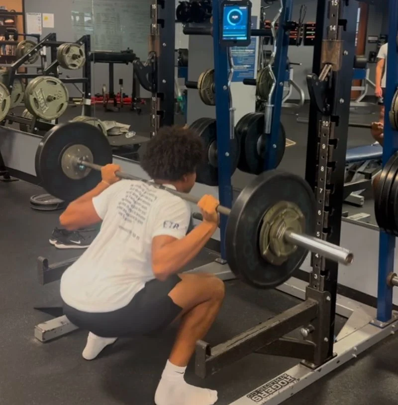 A person is performing a barbell squat in a gym, facing a squat rack. They are wearing a white T-shirt, black shorts, and white socks. Various gym equipment and weight plates are visible in the background.