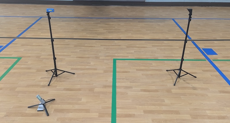 A gym floor with blue and green court lines. Two tall tripods stand on either side and a smaller, flat tripod lies on the ground between them.