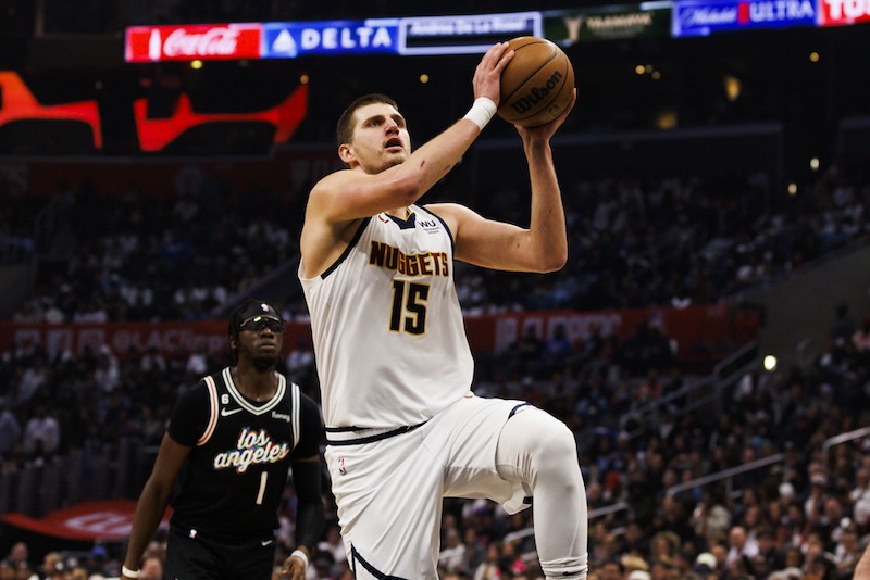A basketball player in a white Nuggets uniform, wearing number 15, is jumping towards the basket with the ball. Another player in a black Los Angeles uniform watches from behind on a crowded court.