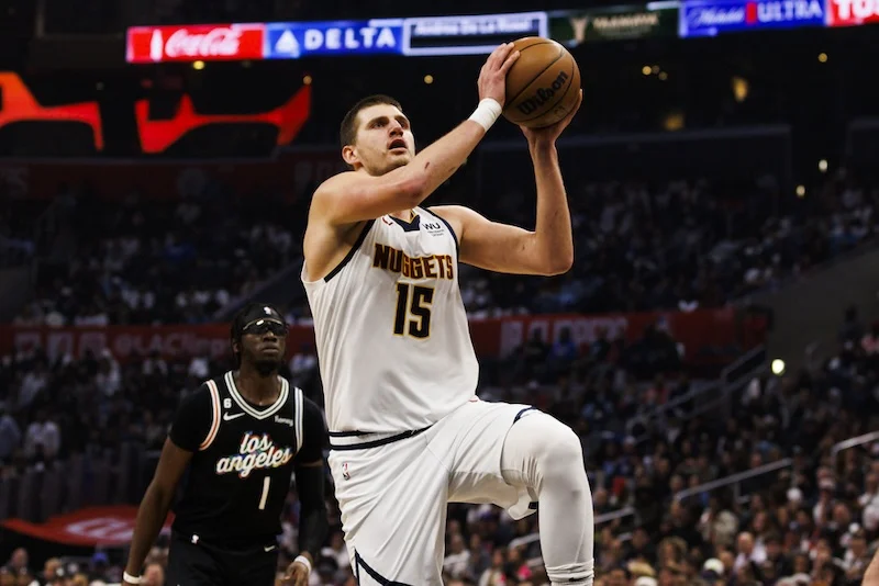 A basketball player in a white Nuggets uniform, wearing number 15, is jumping towards the basket with the ball. Another player in a black Los Angeles uniform watches from behind on a crowded court.