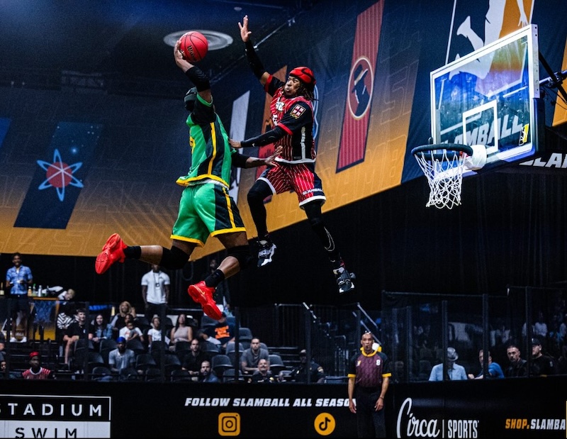 Two athletes in mid-air during a slam dunk in a brightly lit indoor basketball arena. One wears green and yellow, the other red and black. Spectators are visible in the background.
