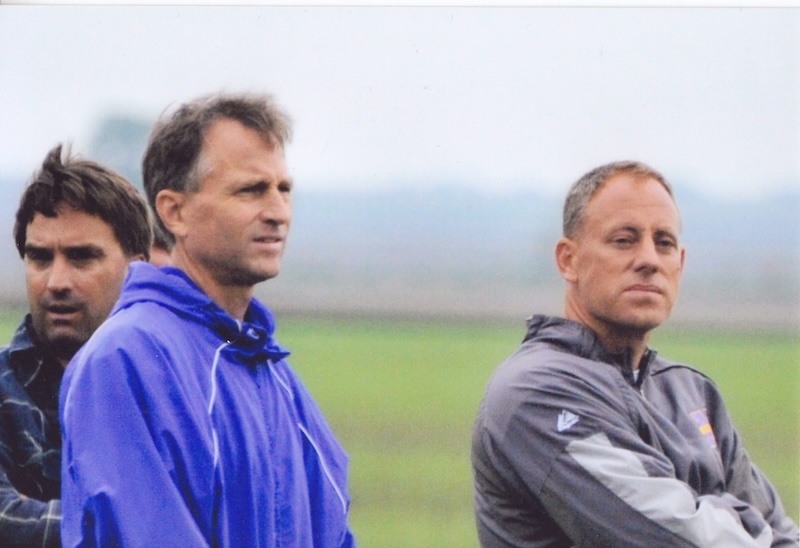Three men stand outdoors, two in blue jackets and one in a gray jacket. They appear to be watching something off-camera. The background is a blurred landscape with cloudy skies.
