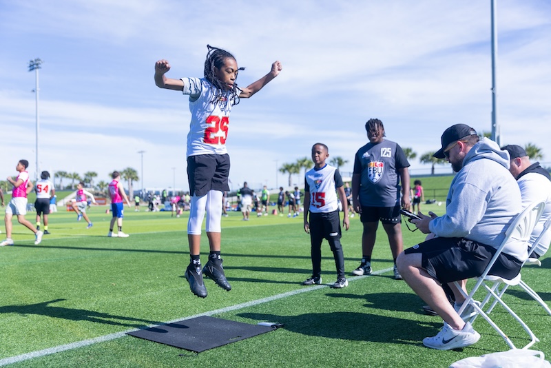 A young athlete wearing a sports jersey and shorts jumps on a grassy field during a sports event. Other participants and a coach, seated and wearing a cap, watch and take notes. Palm trees and a blue sky are in the background.