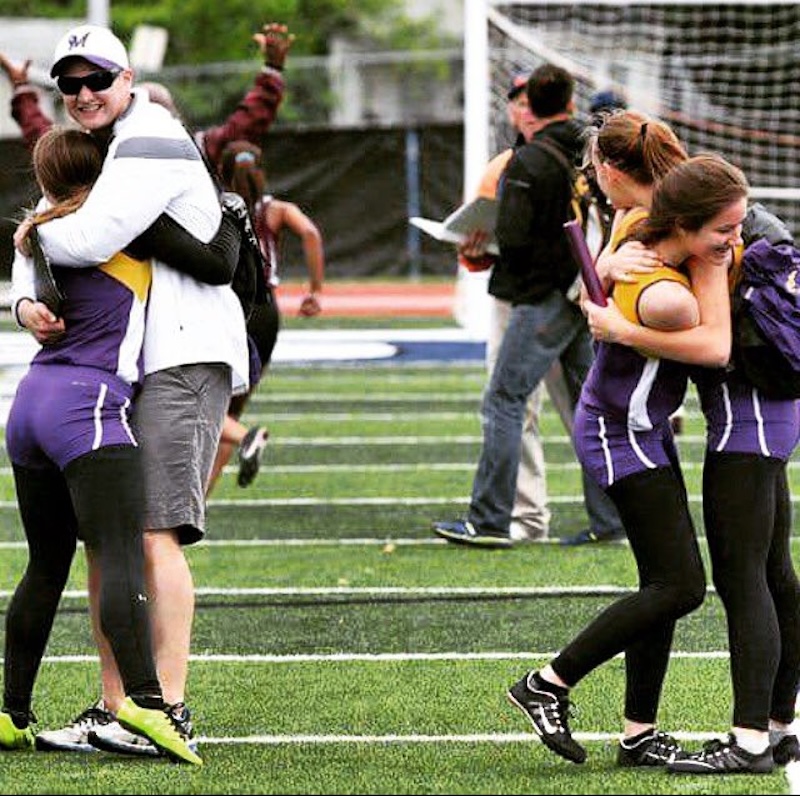 Athletes in purple and yellow uniforms celebrate on a track field. A man in a white jacket embraces one runner, while two other athletes hug each other. In the background, people cheer and walk on the field.
