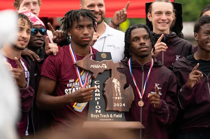 A group of athletes in track uniforms gather around a wooden championship plaque reading 2024 Boys Track & Field Division 1 Champion. Two athletes in the front wear medals, and all appear to be celebrating.