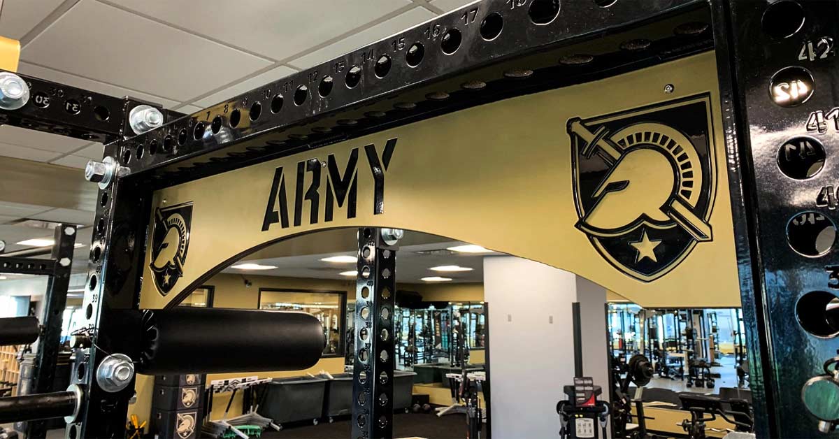Army Football Weight Room|Turf Racks|Rack Tendo|A gym machine with a black frame and seat belt-like strap is positioned on dark carpeted flooring. The machine includes a handle