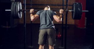 Back Squat|Squat Debate|Bilateral Unilateral Squats|Squatty Non-Squatty|A coach assists a person squatting with weights in a gym. The image compares pros and cons of bilateral versus unilateral squatting for athletes. Text highlights the benefits and limitations of back squats for strength and connective tissue improvement.