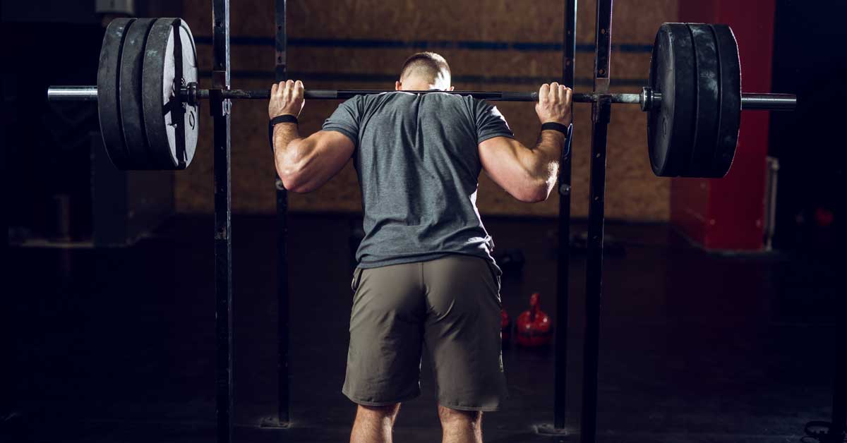 Back Squat|Squat Debate|Bilateral Unilateral Squats|Squatty Non-Squatty|A coach assists a person squatting with weights in a gym. The image compares pros and cons of bilateral versus unilateral squatting for athletes. Text highlights the benefits and limitations of back squats for strength and connective tissue improvement.