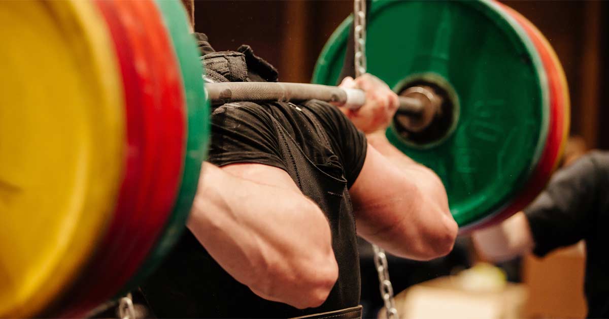 Barbell Squat|Squatting Screen|Barbell Squat|A therapist assists an older man in a physical exercise. The man is squatting in front of a multicolored exercise structure in a room. The therapist guides him by placing a hand on his back for support.