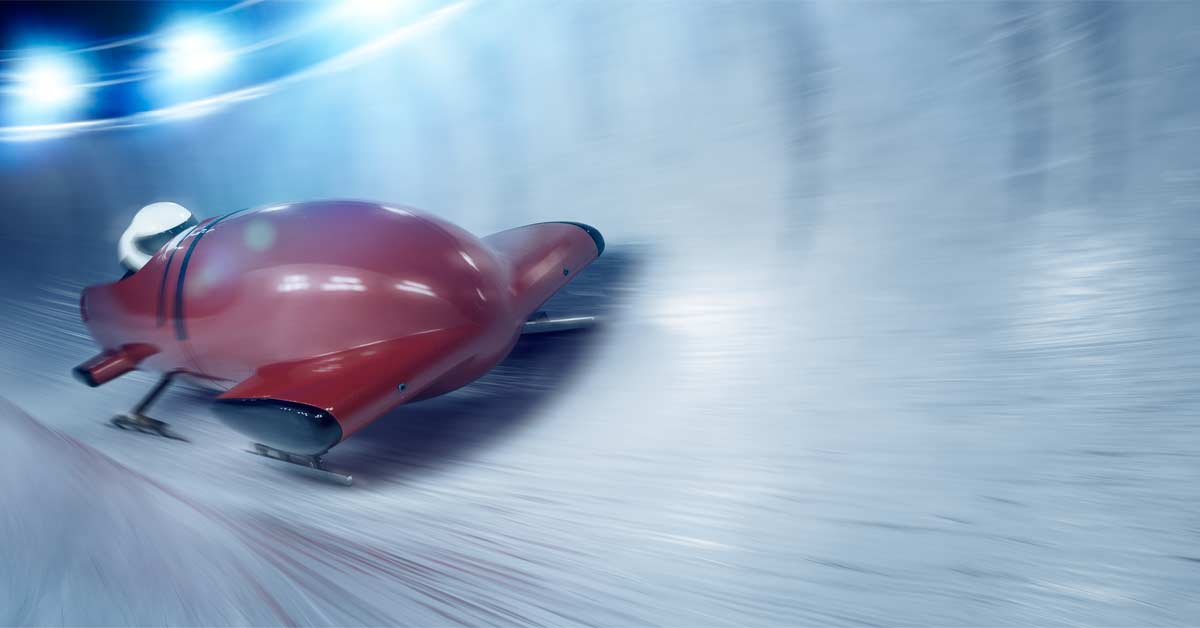 Bobsled|Two men in yellow jackets stand smiling on a bobsled track indoors. They are each holding part of the sled. The track and facility have industrial features