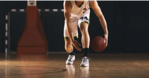 A basketball player in a white jersey dribbles a ball on an indoor court. Focus is on the players lower body and the ball