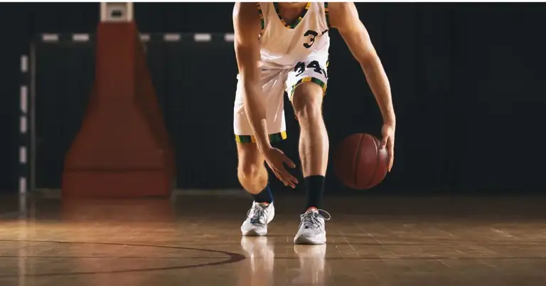A basketball player in a white jersey dribbles a ball on an indoor court. Focus is on the players lower body and the ball