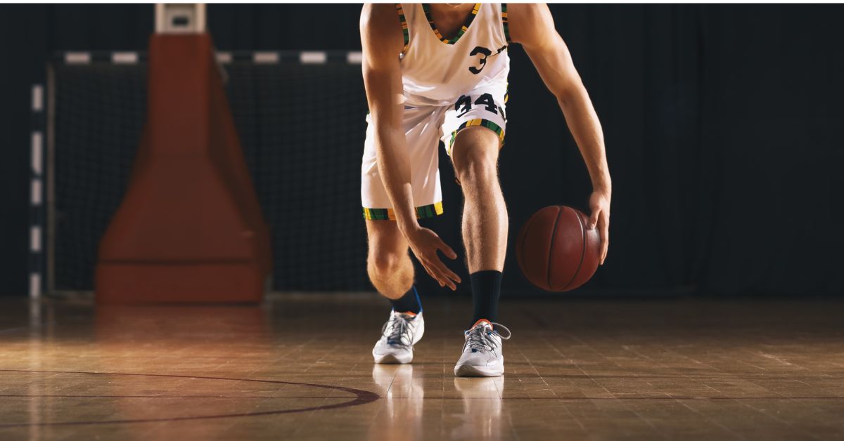 A basketball player in a white jersey dribbles a ball on an indoor court. Focus is on the players lower body and the ball