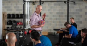 Zac Cupples Speaking|PT Session|A physical therapist assists an athlete in a red shirt during rehabilitation exercises in a room with medical equipment. The athlete wears a knee brace and they appear focused on their movement.