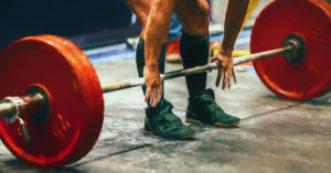 Deadlift Barbell and Weights|Steve Desitter and Barry Ross|Barry Ross Project Day One|Barry Project Plyometrics|A person is running on a red track with a grassy area in the foreground. Two small cones are placed on the track. Behind the track