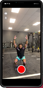 A person in a gym kneeling on a mat with arms raised overhead, positioned in front of a smartphone displaying fitness tracking markers on their joints. The gym has equipment like weights and exercise machines in the background.