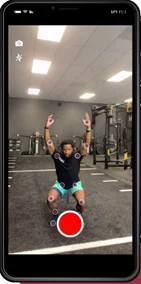 A person in a gym kneeling on a mat with arms raised overhead, positioned in front of a smartphone displaying fitness tracking markers on their joints. The gym has equipment like weights and exercise machines in the background.