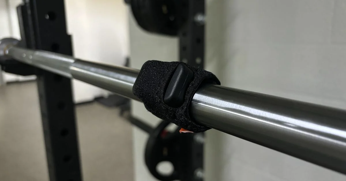 The Enode Pro sensor inside its barbell sleeve attached to a barbell.|An athlete using the Enode sensor on the barbell during squats.|An open black zippered case with foam cutouts and a mesh pocket is shown. Below are a black wristband with an orange and white logo
