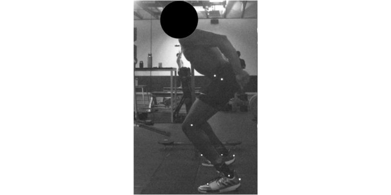 A person in athletic attire is performing a physical activity in a gym setting. The background displays gym equipment, and the persons body is marked with dots, possibly for motion capture analysis. Their face is obscured.