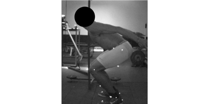A person in a gym performing a squat with feet shoulder-width apart, arms extended backward. They are wearing athletic shorts and shoes. The gym equipment is visible in the background. The face is obscured for privacy.