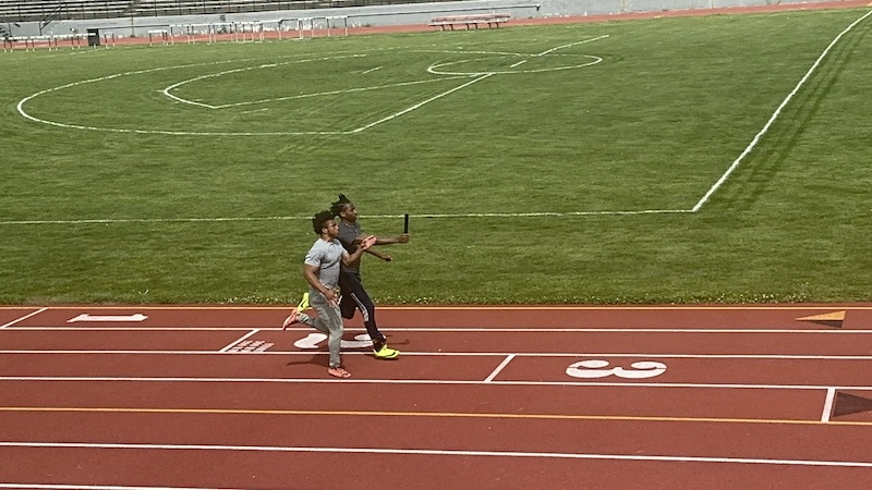 Two athletes are running side by side on a red track, each holding a baton. The grassy field beside the track is visible in the background. The runners appear to be in a relay race.