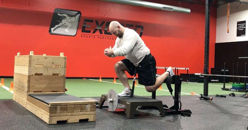 Flywheel Training|A person in an orange shirt and black shorts performs a reverse squat on a platform in a gym. The background features fitness equipment