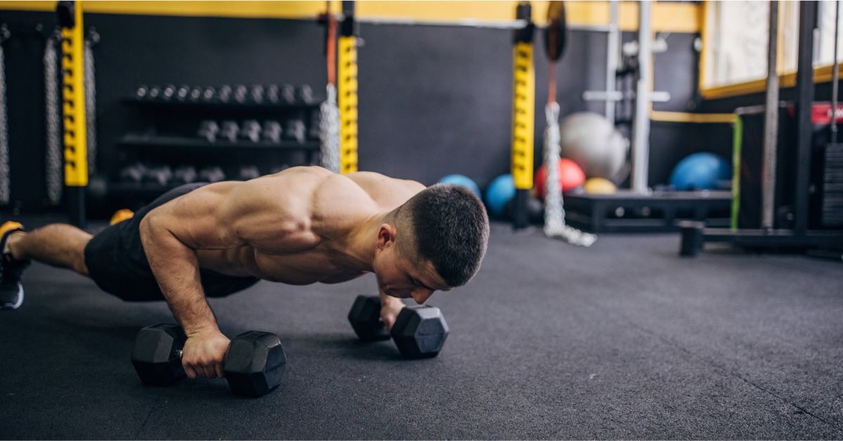 A man in athletic shorts performs a push-up while gripping dumbbells on a gym floor