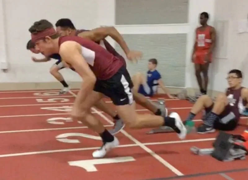 Athletes in maroon and black uniforms spring off starting blocks in an indoor track race. The track is red with white lane markings. A man in a red outfit and other people are visible in the background.
